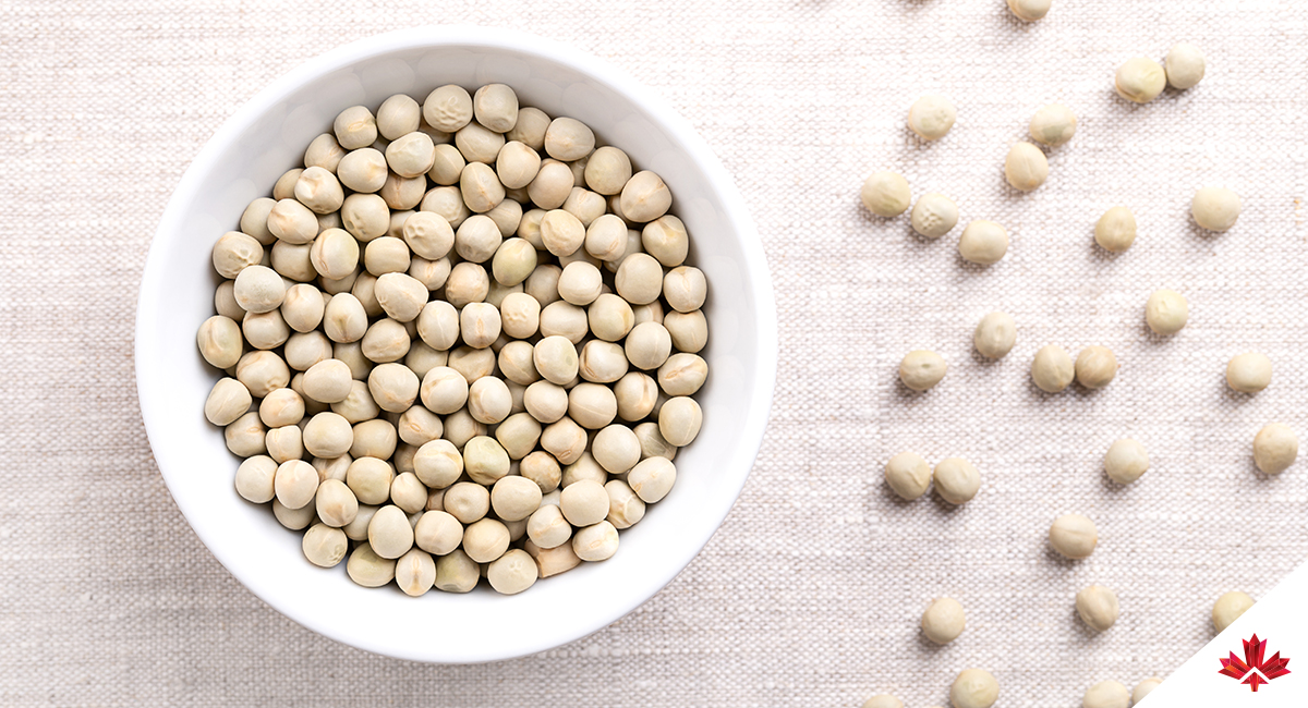 A white bowl filled with dried green peas on a linen surface, with scattered peas around the bowl.