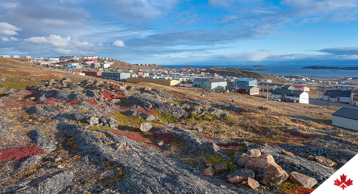 A panoramic view of Iqaluit, Nunavut’s capital, with rocky terrain and sparse vegetation in the foreground. Colourful buildings dot the midground, nestled among rolling hills under a partly cloudy sky. In the distance, Frobisher Bay stretches across the horizon, reflecting the city’s role as a cultural and administrative hub in the Arctic.