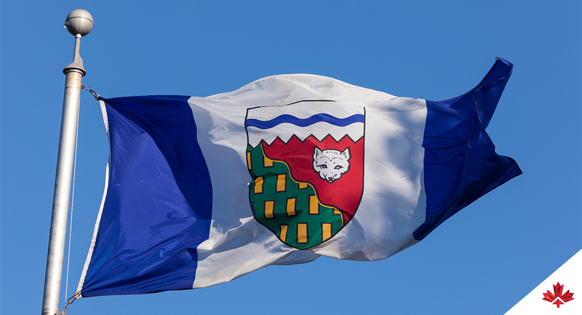 Image of the Northwest Territories flag flying against a clear sky. The flag displays two deep blue side panels and a wide white central panel representing the water, ice and snow of the North. In the centre is the territorial shield, topped by a white section crossed with a wavy blue line symbolizing the polar ice pack and the Northwest Passage. A diagonal line separates a green area with gold rectangles, signifying forested regions and mineral wealth, and a red area with a white Arctic fox, a nod to the region’s historic trade heritage. Together, these visual elements reflect the geography and northern character of the Northwest Territories. 