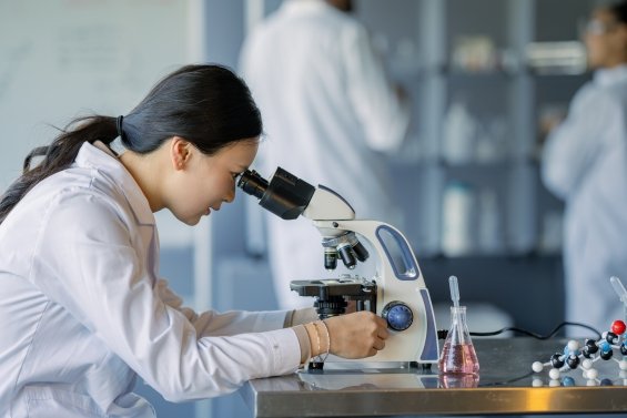 Laboratory technician examining liquid sample in vial, wearing blue gloves, with scientific equipment in background
