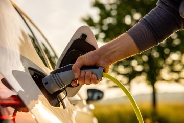 Hand inserting a power cord into a white electric car for charging ecofriendly in a nature landscape on a sunny day.