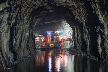 A red heavy‑load vehicle sits in a dim underground tunnel, with bright lights reflecting off the wet, rocky floor. 