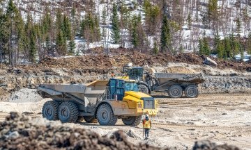 Two dump trucks drive along a dirt mining road, hauling sand past piles of rock, while a worker in an orange vest stands nearby with snowy trees and hills in the background.