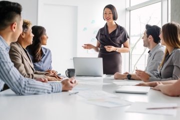 Mature Asian woman leads meeting of multiracial work colleagues discussing ideas and business together sitting at table in modern creative office interior daytime, staff meeting.