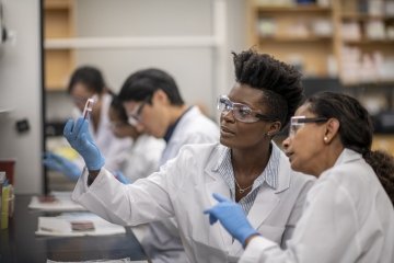 A group of multi-ethnic lab technicians working at desk, each reviewing various specimens closely in cell-culture dishes and discuss findings. Proper safety equipment and attire are being used.