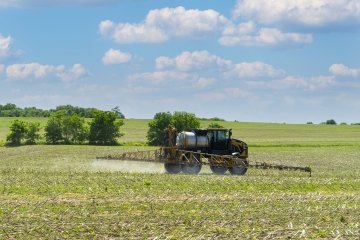 A tractor drives through a green field, spraying fertilizer from a mounted sprayer under a bright sky with scattered clouds. 