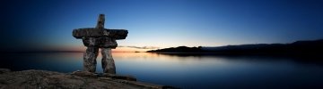 A large stone Inukshuk stands prominently in the foreground, silhouetted against a serene Arctic landscape at sunset. The sky transitions from deep blue to warm orange hues near the horizon, reflecting off calm waters. Distant hills frame the horizon, evoking the vastness and tranquility of Nunavut.