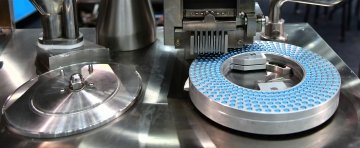 Close‑up of blue capsules arranged in a circular pattern on metallic machinery in a pharmaceutical production line.
