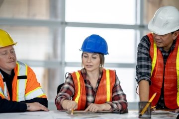 A forewoman wearing safety gear holds a clipboard and looks up while standing at a mining site.