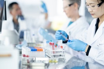 A medical lab technician is seen sorting plasma samples as a small team works together to analyze the samples. They are each wearing lab coats, protective eyewear and gloves as they work diligently.