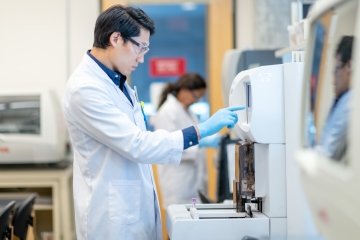 Asian male chemist is working in a laboratory. He is standing in front of a large machine. The medical researcher is preparing to load a sample into the machine for analysis. He is wearing protective gloves and a lab coat. Another scientist is visible working in the background.