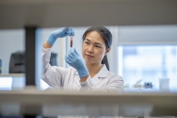 A female lab technician of Asian decent, holds up a vial of blood and looks at it closely before putting it into the centrifuge for processing.