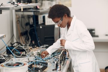 An African American woman scientist works at a lab bench with electronic instruments, including wires, tools, and a microscope.