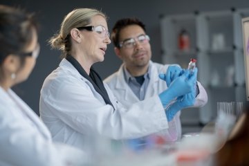 A small team of medical lab staff are seen working together on a vaccine. They are each dressed in white lab coats and personal protective equipment as they discuss and research various elements of the vaccine.