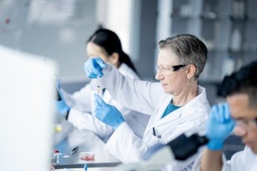 Three laboratory technicians work together in a science lab as they conduct various experiments and record their findings. They are each wearing white lab coats, gloves and protective eyewear as they focus on their work.