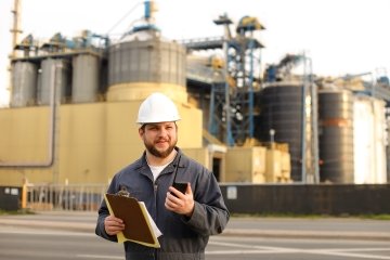 Engineer holding VHF walkie talkie and notes, factory in background. Man wearing helmet and work jumpsuit. Concept of engineering profession and modern technology, worker.