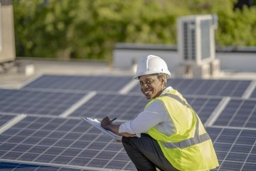 African American female inspecting a rooftop of solar panels to ensure efficiency and function while wearing a hard hat and reflective vest.