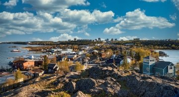 A panoramic view of Yellowknife shows homes, docks and shoreline buildings along Great Slave Lake. Trees display gold and amber tones, while rocky foreground ridges lead toward a distant skyline under bright blue sky and scattered clouds, capturing the vastness and character of the Northwest Territories.